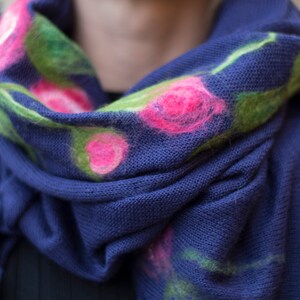 May include: A blue knitted scarf with pink and green floral embroidery. The scarf is folded and draped over a person's shoulders.
