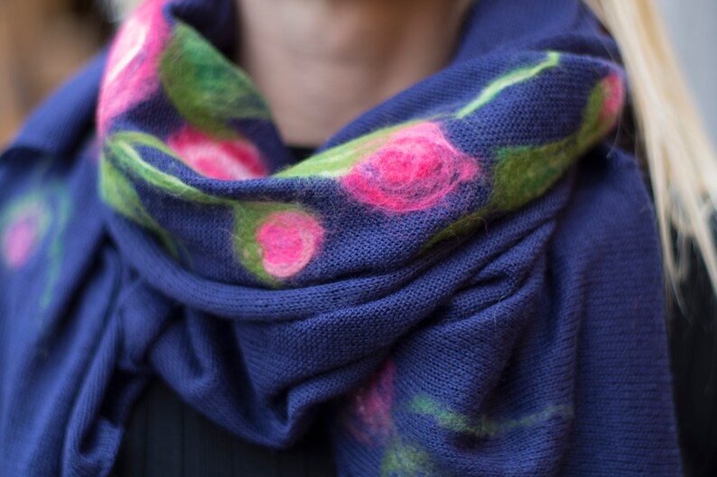 May include: A blue knitted scarf with pink and green floral embroidery. The scarf is folded and draped over a person's shoulders.