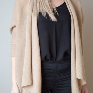 Puede incluir: Un cárdigan beige con un frente drapeado y mangas largas. El cárdigan se lleva sobre una blusa negra y pantalones negros.