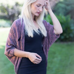 May include: A woman with long blonde hair is wearing a pink and gray cardigan over a black dress. She is standing in a grassy area with a blurred background.
