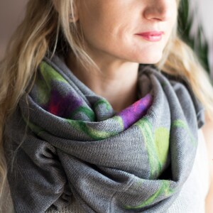 May include: A gray scarf with a floral pattern in shades of green, purple, and pink. The scarf is made of a soft, knitted fabric and is worn around the neck.