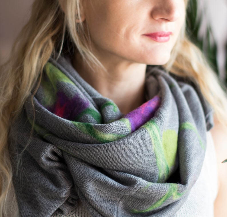 May include: A gray scarf with a floral pattern in shades of green, purple, and pink. The scarf is made of a soft, knitted fabric and is worn around the neck.