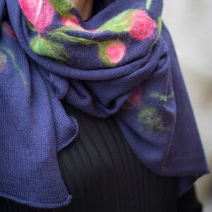 May include: A blue knitted scarf with a floral design featuring pink and green flowers. The scarf is folded and draped around a person's neck.