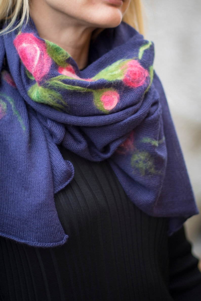May include: A blue knitted scarf with a floral design featuring pink and green flowers. The scarf is folded and draped around a person's neck.