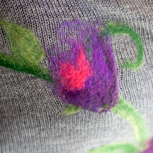 May include: A close-up of a gray knitted fabric with a purple and pink flower design. The flower is embroidered with wool yarn and has a green stem.