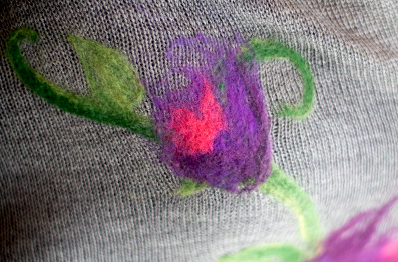 May include: A close-up of a gray knitted fabric with a purple and pink flower design. The flower is embroidered with wool yarn and has a green stem.