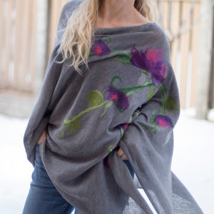 May include: A gray knitted poncho with a floral design featuring purple, pink, and green flowers. The poncho is draped over a person's shoulders and is worn with blue jeans.
