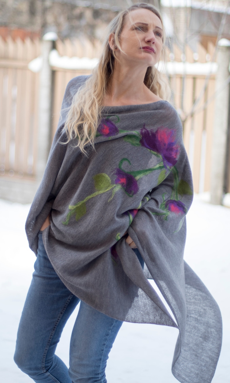 May include: A gray knitted poncho with a floral design featuring purple, pink, and green flowers. The poncho is draped over a person's shoulders and is worn with blue jeans.