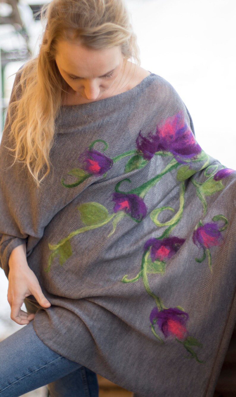May include: A gray knitted poncho with a floral design featuring purple, pink, and green flowers. The poncho is made of a soft, lightweight fabric and has a relaxed fit.