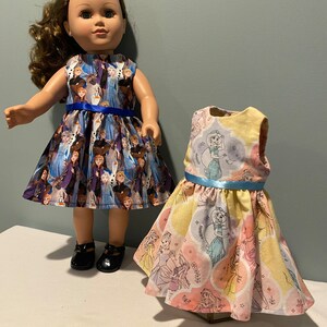 May include: Two doll dresses, one with a blue and white Frozen print and the other with a yellow and pink princess print. Both dresses have blue sashes.