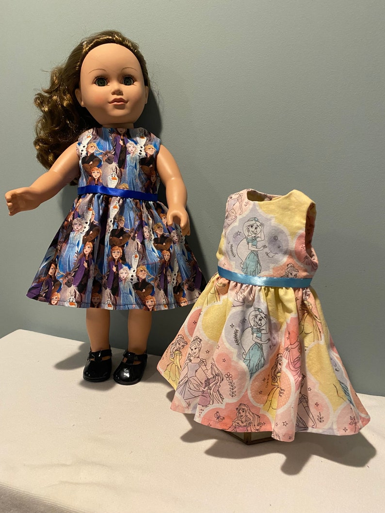 May include: Two doll dresses, one with a blue and white Frozen print and the other with a yellow and pink princess print. Both dresses have blue sashes.