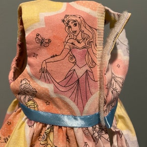 May include: A sleeveless dress for a doll, featuring a colorful print of Sleeping Beauty and other Disney princesses. The dress has a blue ribbon around the waist and a zipper closure on the back.
