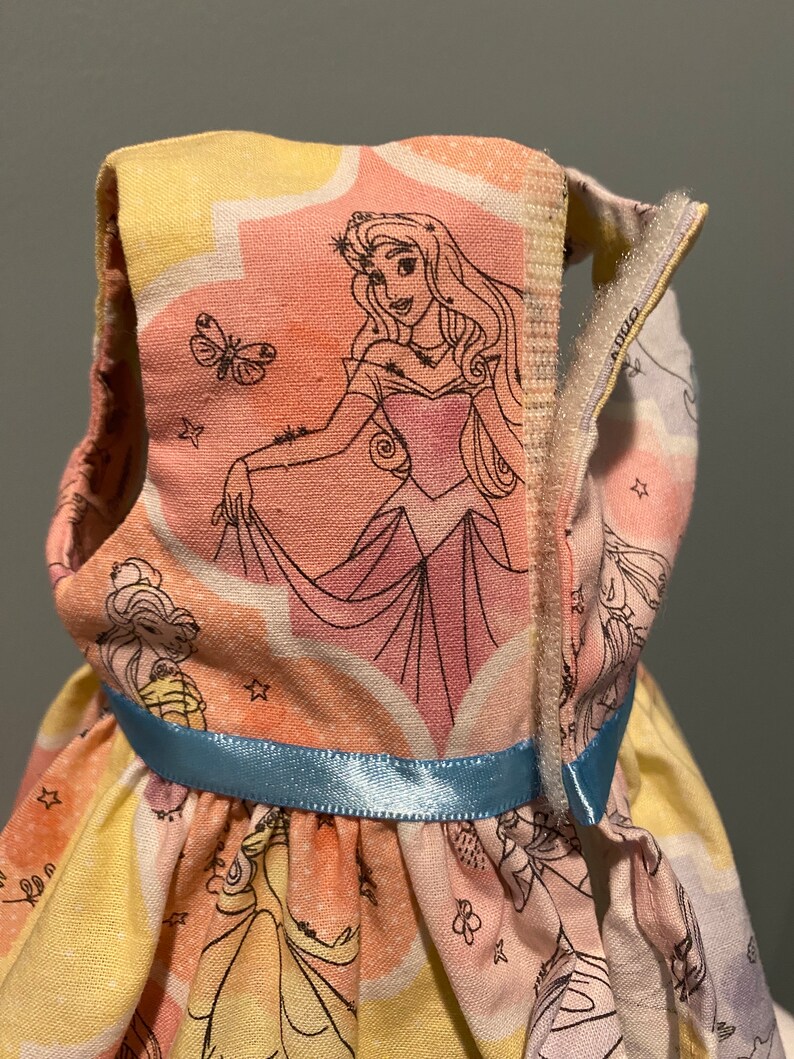 May include: A sleeveless dress for a doll, featuring a colorful print of Sleeping Beauty and other Disney princesses. The dress has a blue ribbon around the waist and a zipper closure on the back.
