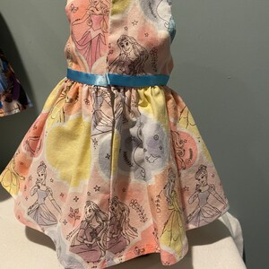 May include: A yellow, pink, and blue sleeveless dress with a light blue ribbon. The dress has a pattern of Disney princesses, including Cinderella, Rapunzel, and Aurora.