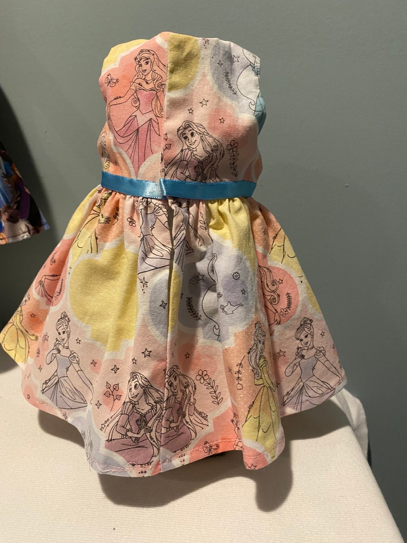 May include: A yellow, pink, and blue sleeveless dress with a light blue ribbon. The dress has a pattern of Disney princesses, including Cinderella, Rapunzel, and Aurora.