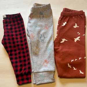 May include: Three pairs of children's leggings. One pair is red and black plaid, one pair is gray with a robot pattern, and one pair is rust with a white bird pattern.