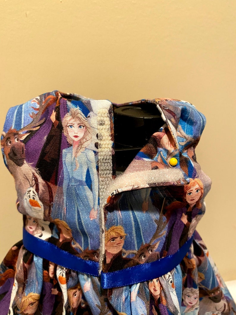 May include: A blue and purple dress with a blue ribbon. The dress is covered in images of characters from the movie Frozen II.