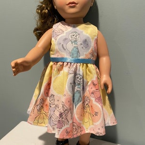 May include: A doll wearing a yellow, pink, and blue dress with a print of Disney princesses. The dress has a blue ribbon around the waist. The doll is wearing black shoes.