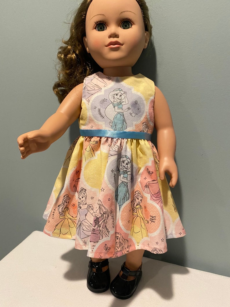 May include: A doll wearing a yellow, pink, and blue dress with a print of Disney princesses. The dress has a blue ribbon around the waist. The doll is wearing black shoes.