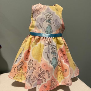 May include: A yellow sleeveless dress with a blue ribbon belt. The dress is printed with images of Disney princesses in pastel colors.