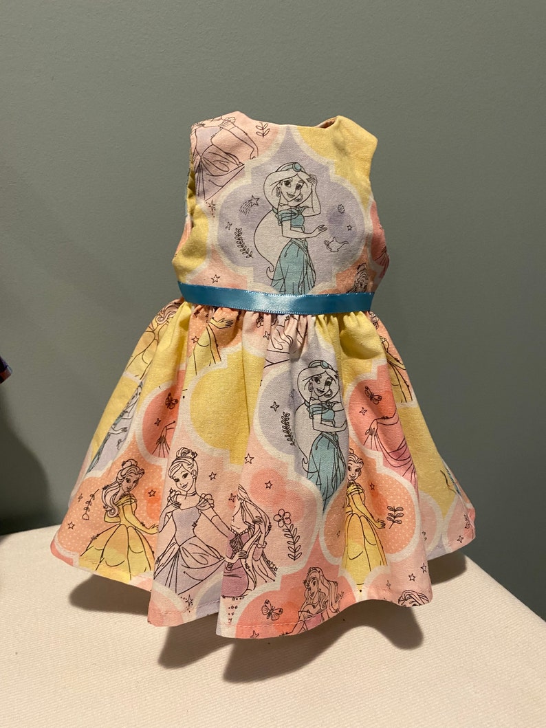May include: A yellow sleeveless dress with a blue ribbon belt. The dress is printed with images of Disney princesses in pastel colors.