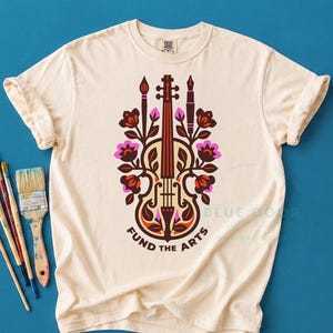 May include: A cream-colored t-shirt featuring a central graphic of a violin integrated with floral motifs, a paintbrush, and a fountain pen. The text "FUND THE ARTS" is printed in a dark brown arc below the illustration. Four paintbrushes are visible.