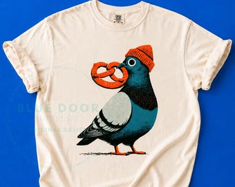 May include: A cream-colored t-shirt featuring a graphic illustration of a blue pigeon wearing an orange beanie and holding a brown pretzel in its beak. The design is centered on the chest of the shirt, which has short sleeves and a crew neckline.