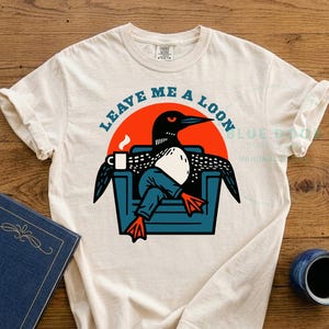Leave Me A Loon graphic tee laid flat on a weathered wooden dock beside a book and steaming camp mug, ivory Comfort Colors 1717 shirt featuring a funny loon illustration relaxing in an armchair, retro bird lover gift for lake house and cabin living