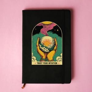 May include: Black journal with a colorful illustration of hands holding a crystal ball. The image features a sun, clouds, and the text "TRUST YOUR INTUITION". The journal has a ribbon bookmark.