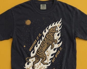 May include: A charcoal gray short-sleeve t-shirt featuring a graphic of a tiger leaping through flames. The text "WHATEVER IT IS. GET AFTER IT." is printed in gold lettering alongside the tiger, with small stars and a moon in the background.