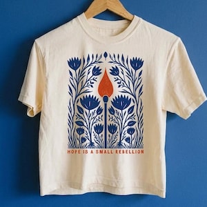 May include: A cream-colored short-sleeve t-shirt featuring a central graphic of a lit match surrounded by blue floral patterns. Below the illustration, the text reads "HOPE IS A SMALL REBELLION" in orange capital letters. The shirt hangs on a wooden hanger.