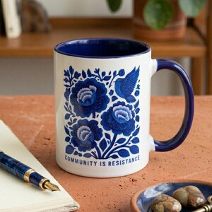 May include: A white ceramic mug with a dark blue interior and handle, featuring a blue floral pattern with a bird and the text "COMMUNITY IS RESISTANCE" printed in blue. The mug sits alongside a blue and gold fountain pen and a small blue dish.