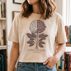 May include: Beige t-shirt with a graphic of a brain-flower and the text "OVERTHINKING in full bloom". The design is in shades of brown and purple. The shirt is paired with blue jeans.