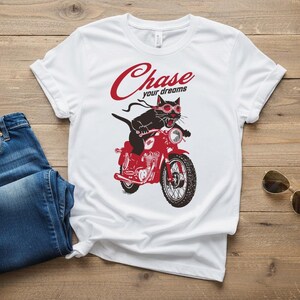 May include: White t-shirt with a graphic of a black cat riding a red motorcycle, wearing goggles. The text "Chase your dreams" is above the cat. Blue jeans and sunglasses are next to the shirt.