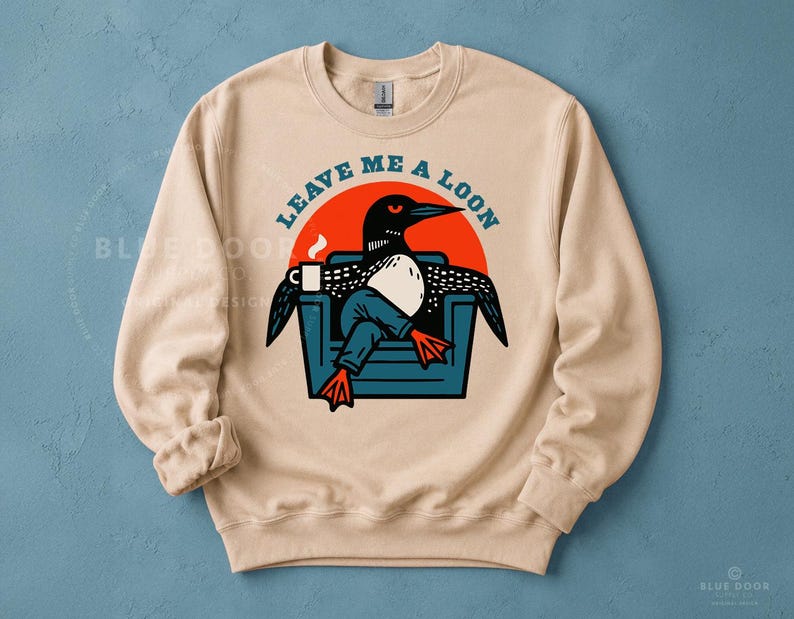 Leave Me A Loon sweatshirt in sand laid flat on blue background, funny loon crewneck featuring a bird lounging in an armchair with coffee, cozy lake gift for Minnesota winters