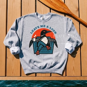 Loon sweatshirt in sport gray hanging on a wooden dock beside a canoe paddle, funny loon crewneck styled for camping and lake life outdoor gifting