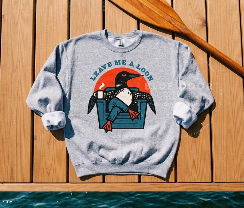 Loon sweatshirt in sport gray hanging on a wooden dock beside a canoe paddle, funny loon crewneck styled for camping and lake life outdoor gifting