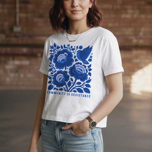 Puede incluir: Una camiseta blanca de manga corta con un estampado gr&aacute;fico azul de flores estilizadas y un p&aacute;jaro. Debajo del dise&ntilde;o floral, el texto "COMMUNITY IS RESISTANCE" aparece en letras may&uacute;sculas azules. Tiene cuello redondo y dobladillo recto.