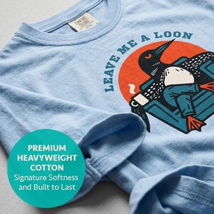 Minnesota loon gift shirt folded neatly in bay blue heavyweight cotton, close-up of the funny loon in an armchair graphic with red sun and coffee mug design, retro bird tee for cabin and lake house gifting