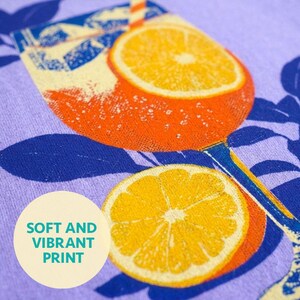 Close-up of a purple fabric with a vibrant orange cocktail glass design, featuring orange slices and a straw. The text "SOFT AND VIBRANT PRINT" is in a circle on the lower left.