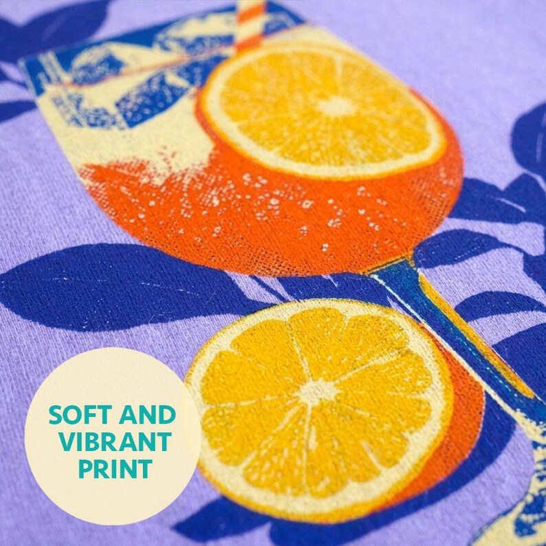 Close-up of a purple fabric with a vibrant orange cocktail glass design, featuring orange slices and a straw. The text "SOFT AND VIBRANT PRINT" is in a circle on the lower left.