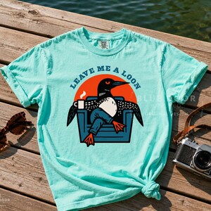 Funny loon tee in island reef laid flat on a sun-bleached dock with vintage sunglasses and a film camera styled beside it, lake life shirt with retro bird graphic, camp-core aesthetic for outdoorsy gift shopping