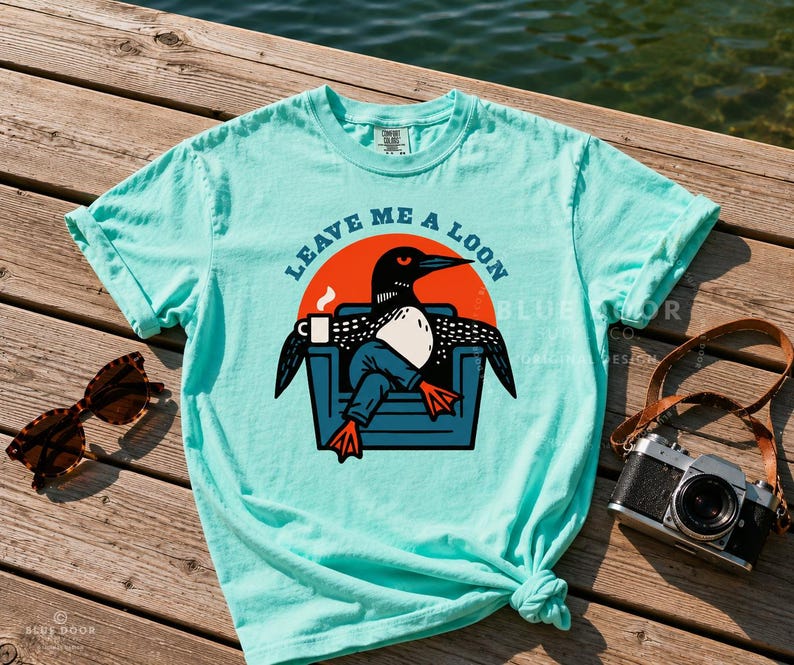 Funny loon tee in island reef laid flat on a sun-bleached dock with vintage sunglasses and a film camera styled beside it, lake life shirt with retro bird graphic, camp-core aesthetic for outdoorsy gift shopping