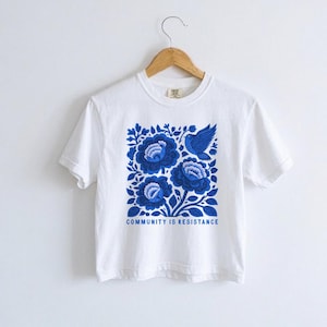 Puede incluir: Una camiseta blanca de manga corta con un gr&aacute;fico cuadrado en tonos azules. El dise&ntilde;o muestra tres grandes flores estilizadas y un p&aacute;jaro rodeado de hojas. Debajo de la ilustraci&oacute;n, el texto dice COMMUNITY IS RESISTANCE.
