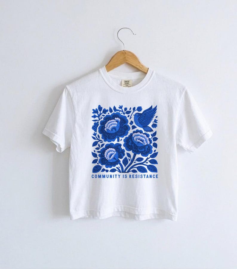 Puede incluir: Una camiseta blanca de manga corta con un gr&aacute;fico cuadrado en tonos azules. El dise&ntilde;o muestra tres grandes flores estilizadas y un p&aacute;jaro rodeado de hojas. Debajo de la ilustraci&oacute;n, el texto dice COMMUNITY IS RESISTANCE.