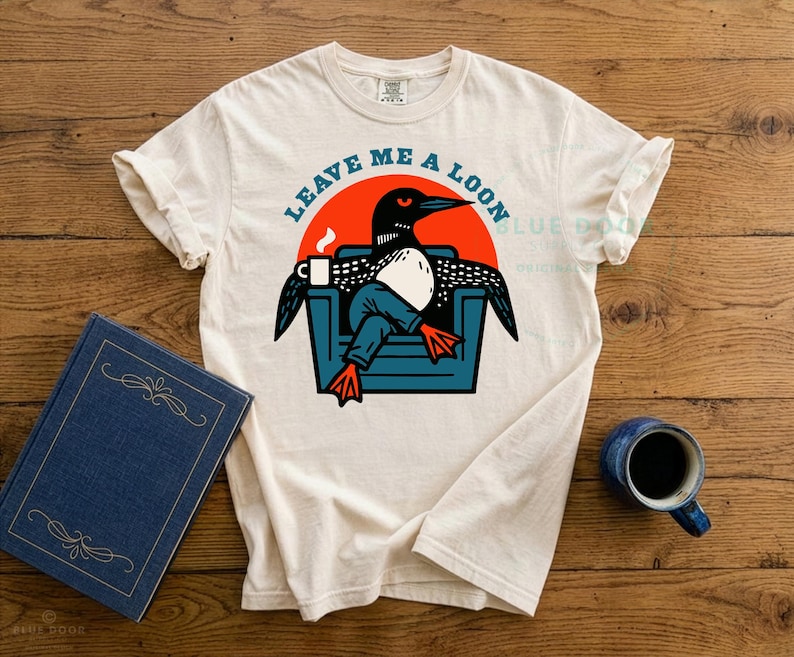 Leave Me A Loon graphic tee laid flat on a weathered wooden dock beside a book and steaming camp mug, ivory Comfort Colors 1717 shirt featuring a funny loon illustration relaxing in an armchair, retro bird lover gift for lake house and cabin living