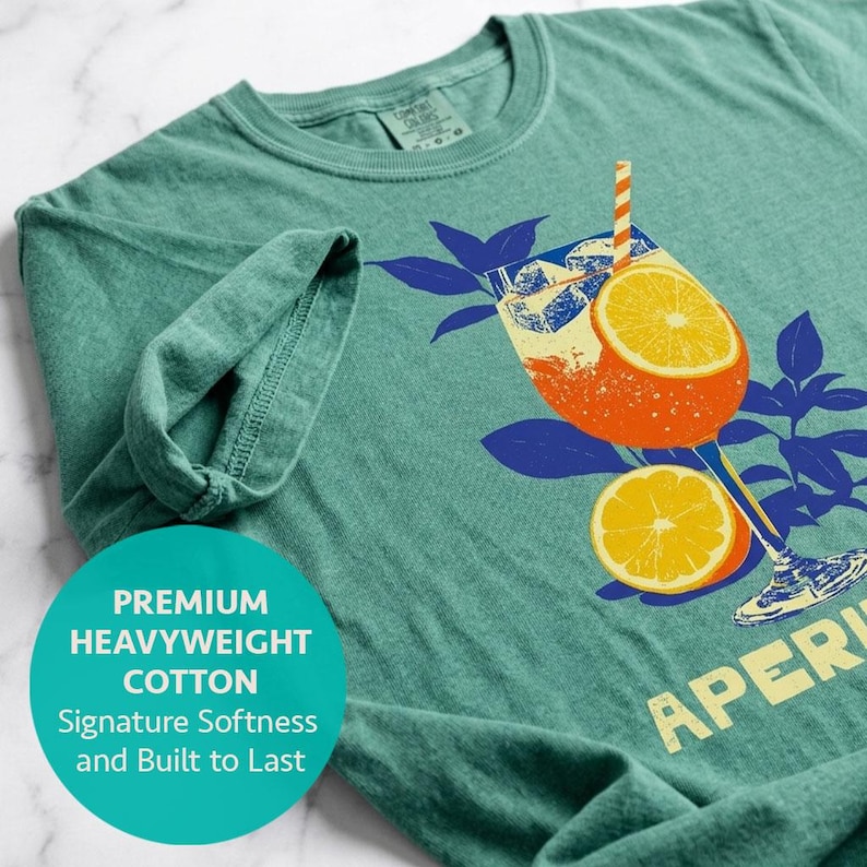 A light green comfort colors t-shirt featuring a graphic of an orange cocktail with ice cubes, orange slices, and a striped straw. The shirt is made of heavyweight cotton.