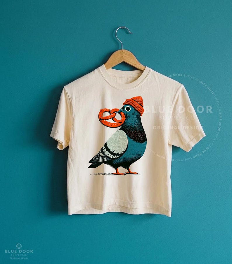 NYC crop top in ivory Comfort Colors cotton on a teal background, a New York pigeon in an orange beanie holding a pretzel