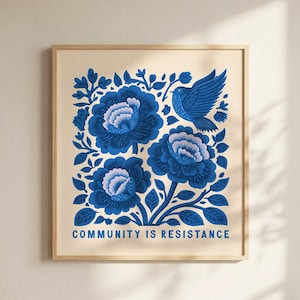 May include: A square framed art print featuring a cream background with a blue floral design and a bird. The illustration includes three large, detailed blue flowers, foliage, and a bird in flight. The text "COMMUNITY IS RESISTANCE" is printed in blue at the bottom.