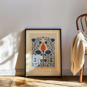 May include: A framed art print featuring a central orange matchstick surrounded by symmetrical blue floral illustrations on a cream background. The text HOPE IS A SMALL REBELLION is printed in orange at the bottom of the rectangular composition.